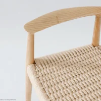 A close-up of a wooden chair with a woven seat and a curved wooden backrest, shown against a plain light background. The craftsmanship of the natural wood and woven pattern is clearly visible.