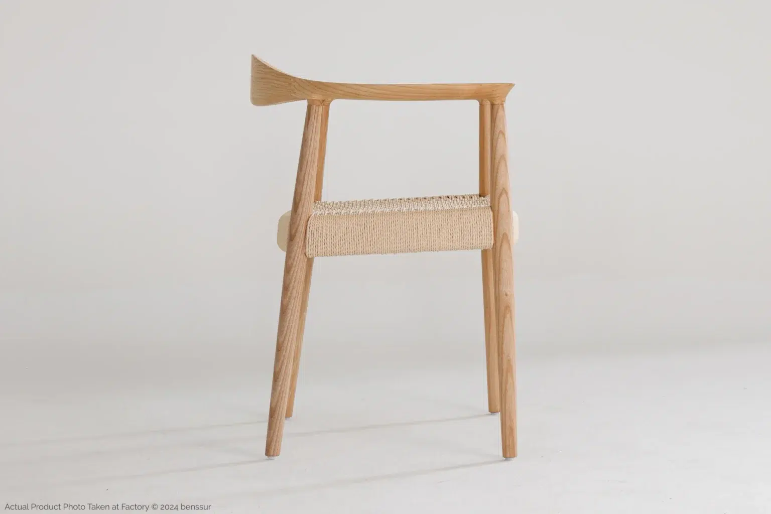 Hans Wegner_Round Chair_Replica_Paper Rope_Natural_Ash_Right A minimalist wooden chair with an asymmetrical backrest and woven seat, set against a plain white background. The design features clean lines and a natural wood finish.