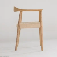 A minimalist wooden chair with an asymmetrical backrest and woven seat, set against a plain white background. The design features clean lines and a natural wood finish.