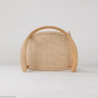 A small woven basket with a curved wooden handle and rounded wooden sides, placed on a white surface against a plain white background.
