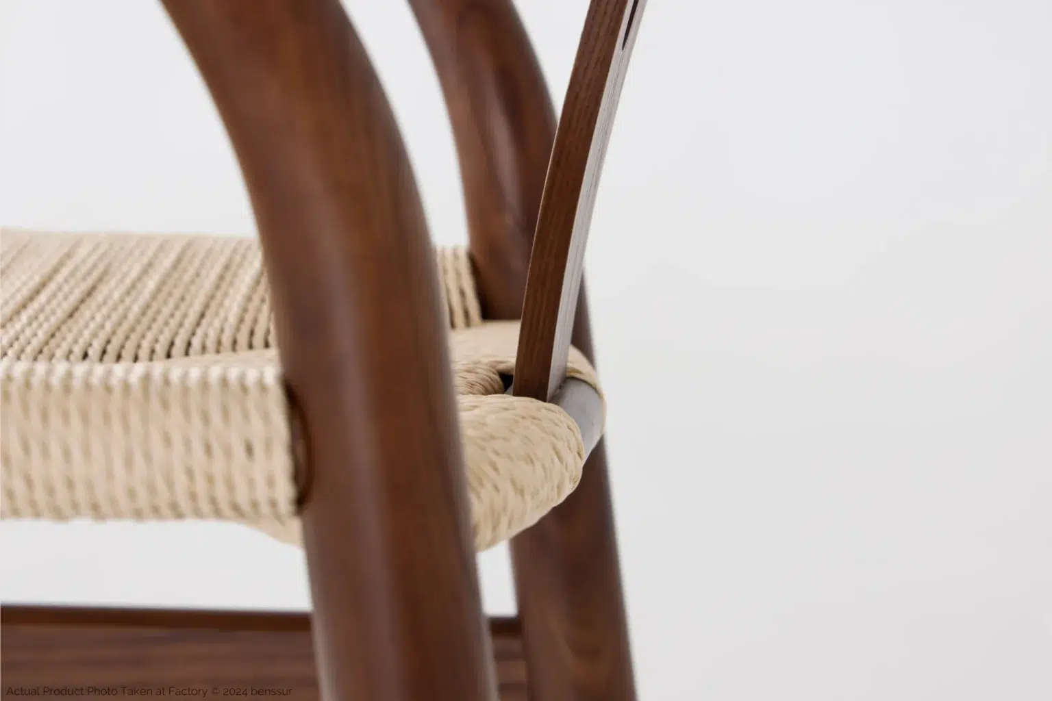 Hans Wegner_Wishbone Chair_Replica_Paper Rope_Dark Brown_Ash_Detail_2 Close-up of a chair with smooth, curved wooden legs and a woven natural fibre seat, set against a plain white background.