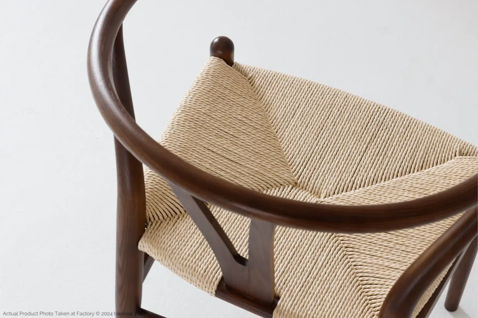 Hans Wegner_Wishbone Chair_Replica_Paper Rope_Dark Brown_Ash_Detail_3 A close-up of a wooden chair with a curved backrest and a woven, light-coloured seat, photographed against a plain white background.
