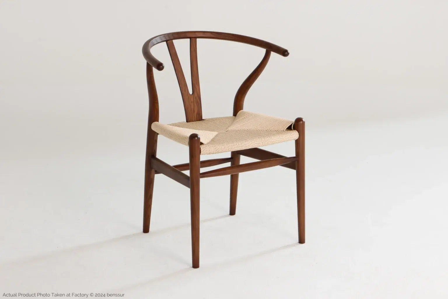 Hans Wegner_Wishbone Chair_Replica_Paper Rope_Dark Brown_Ash_Front Right A wooden Wishbone chair with a curved backrest and woven seat sits on a plain white background. The design is minimalist and elegant, featuring smooth, rounded edges and tapered legs.