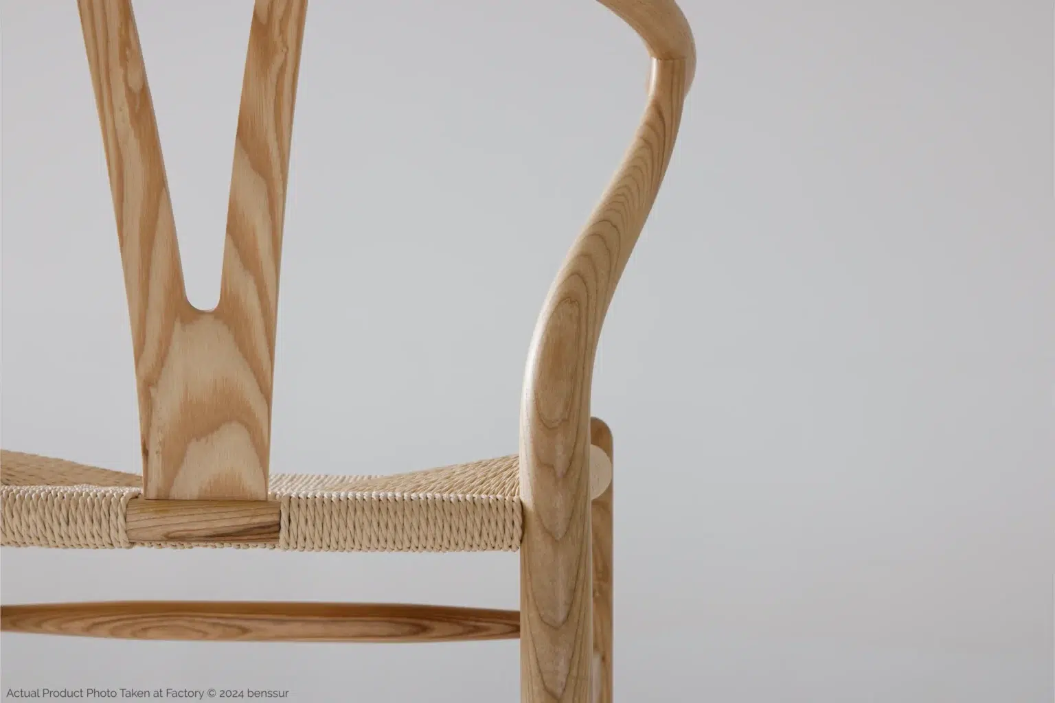 Hans Wegner_Wishbone Chair_Replica_Paper Rope_Natural_Ash_Detail_2 Close-up of a wooden chair with a curved backrest and woven seat, set against a plain light background. Only the right side of the chair is visible, highlighting its smooth finish and elegant design.