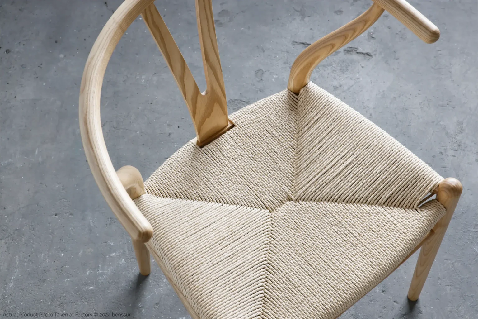 Hans Wegner_Wishbone Chair_Replica_Paper Rope_Natural_Ash_Detail_3 A wooden chair with a curved backrest and woven seat is placed on a grey concrete floor, viewed from above. The natural wood and intricate seat weave create a minimalist, Scandinavian design.