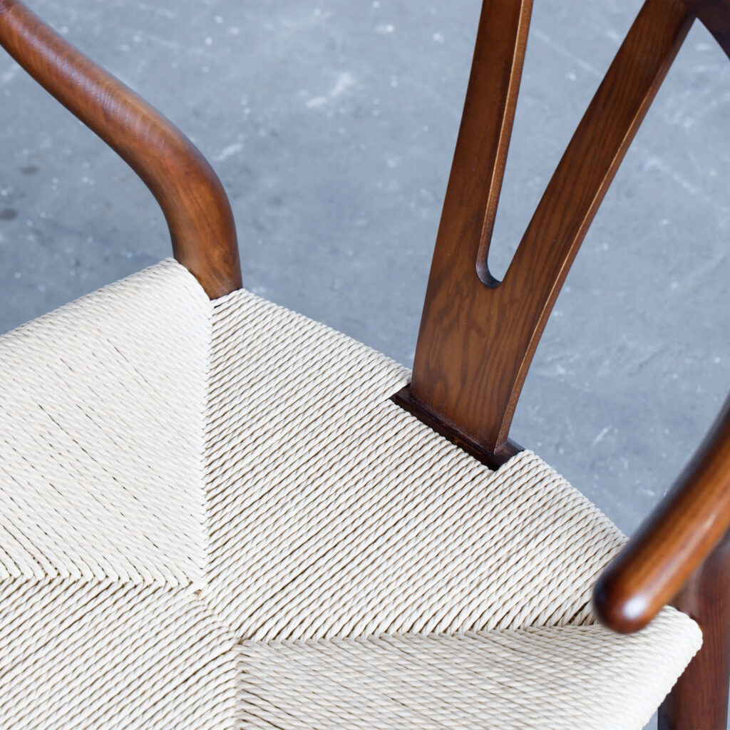 Close-up of a wooden chair with a V-shaped backrest and a woven, light-coloured seat, set against a grey textured floor.