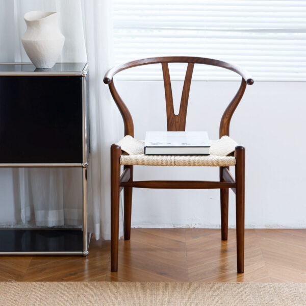 A wooden chair with a woven seat sits next to a black cupboard and white vase, beneath a window with blinds. A closed laptop rests on the chair. The floor features a herringbone wood pattern.