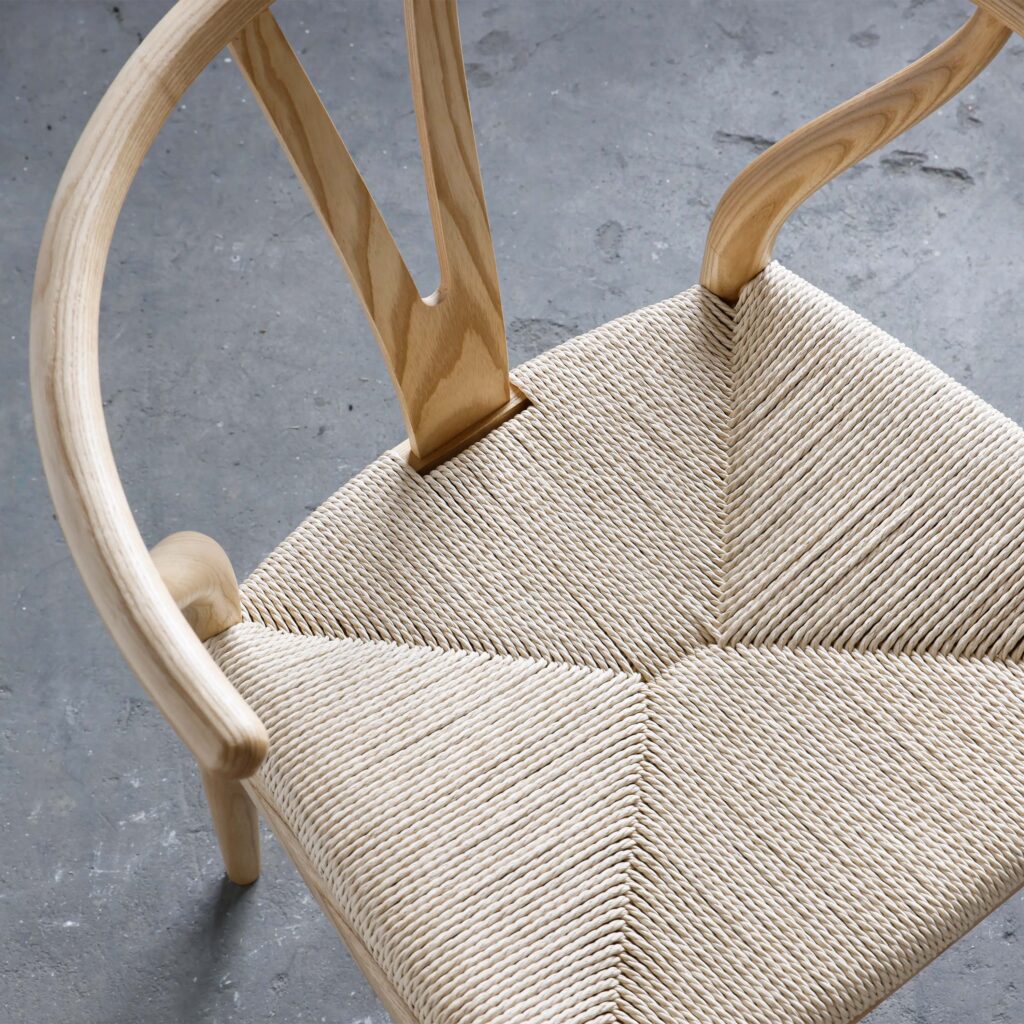 A top-down view of a wooden chair with a curved backrest and a woven seat, placed on a grey concrete floor. The chair features smooth, light-coloured wood and intricate weaving.