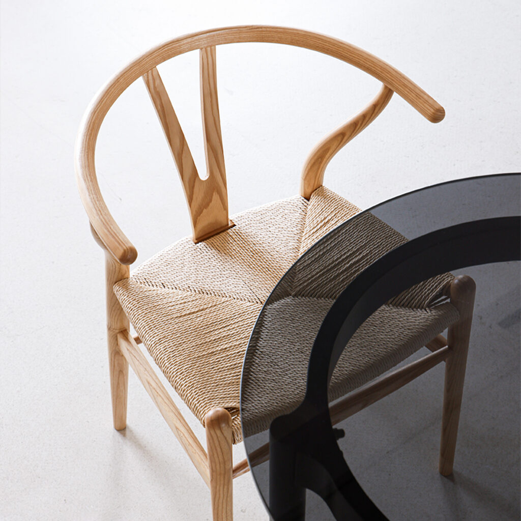 A wooden chair with a curved backrest and woven seat is positioned next to a round, black-tinted glass table on a light-coloured floor.