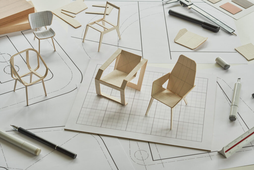 Paper models of chairs sit on architectural blueprints and graph paper, surrounded by pens, a ruler, and design tools, illustrating the process of furniture design and prototyping.