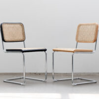 Two modern chairs with chrome cantilever frames, woven cane seats and backs; one chair has a black frame around the seat and back, whilst the other has a light wood frame. They are placed facing each other in a bright room.