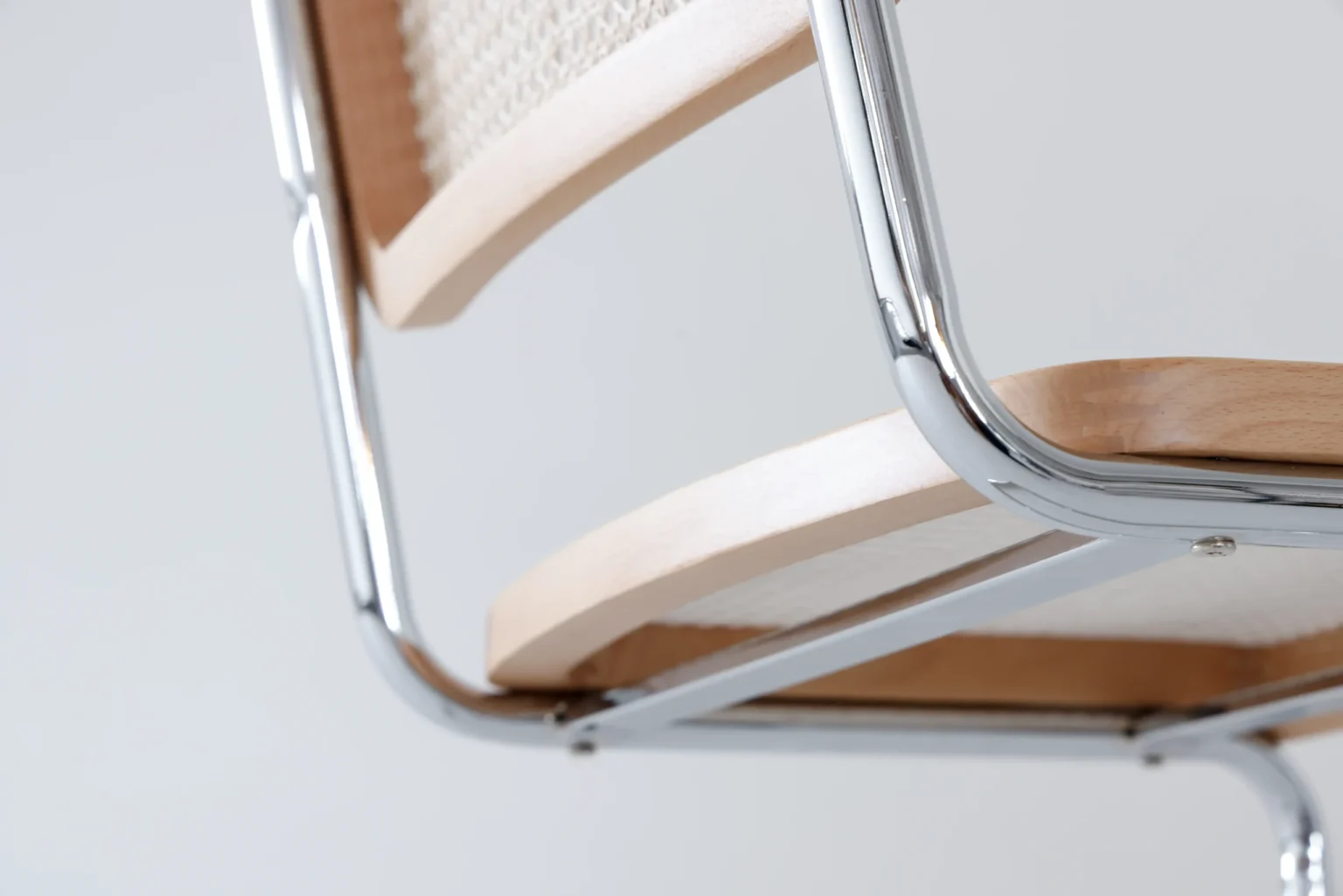 Detail display2 Close-up of a modern chair's frame, showing shiny chrome metal, light wood elements, and beige woven material on the backrest, set against a plain light grey background.