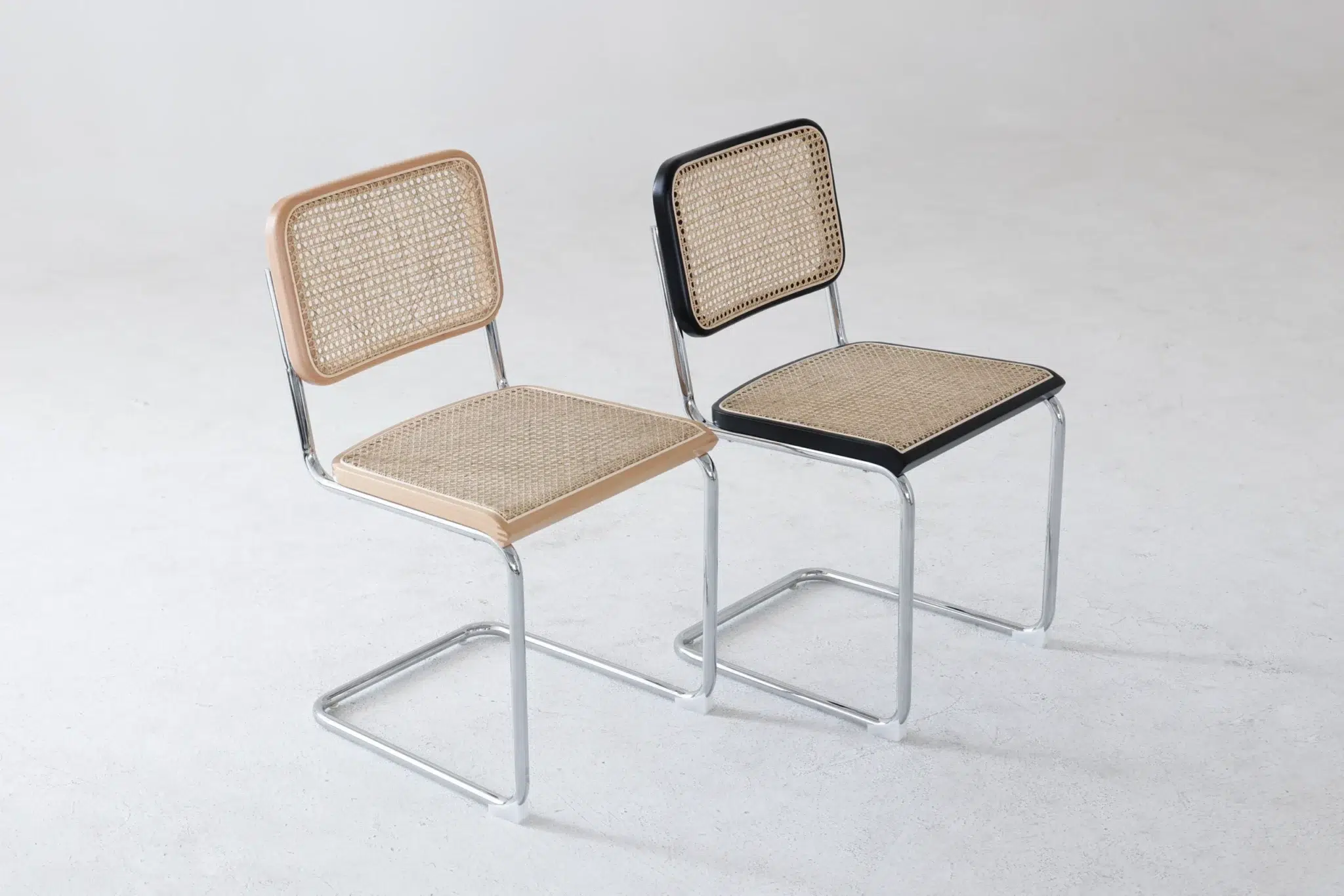 Two modern chairs with woven cane seats and backs, metal tubular frames, and no armrests. One chair has light wood accents; the other has black accents. Both are positioned on a light, plain surface.