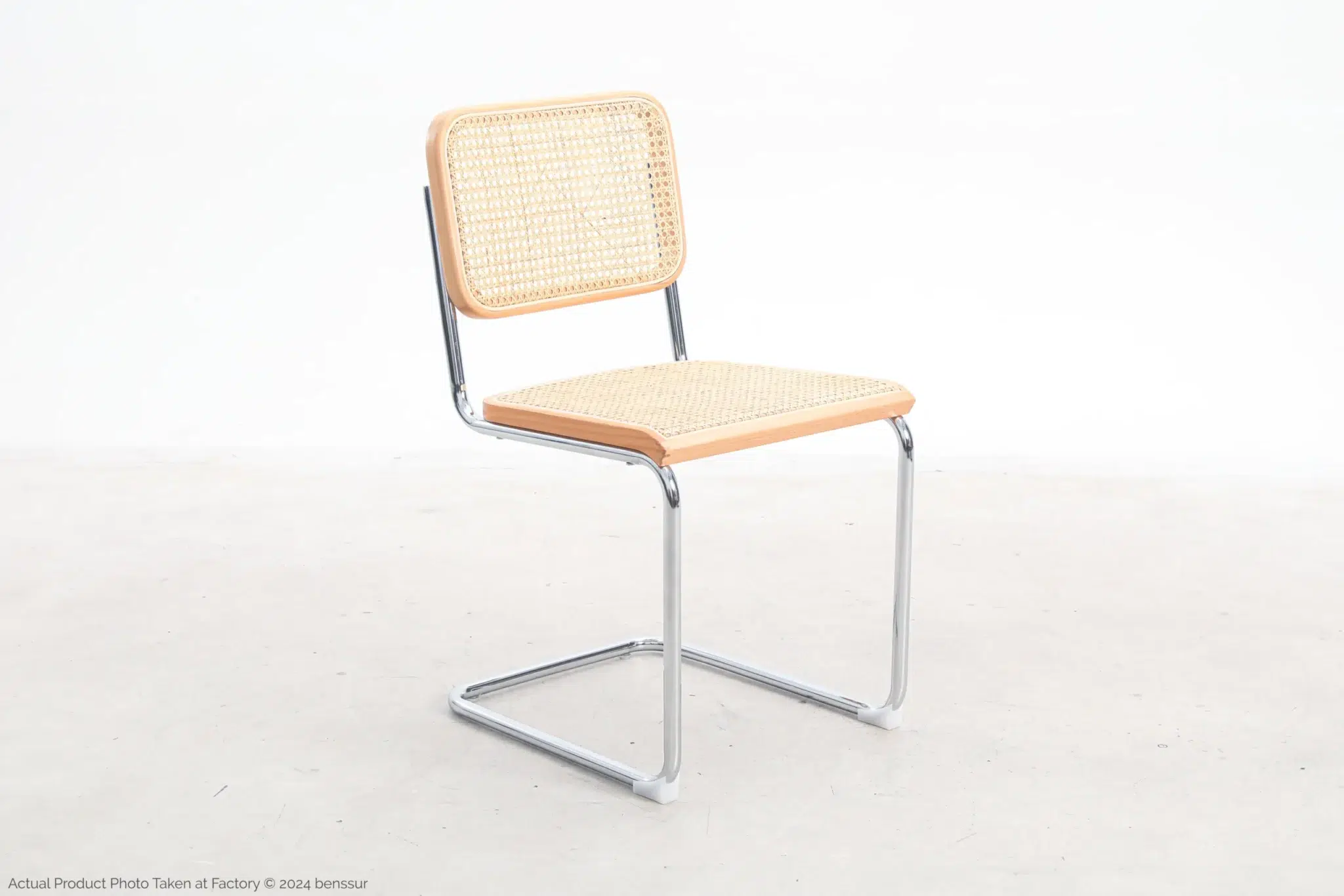 A modern chair with a woven cane backrest and seat, featuring a light wooden frame and a sleek, cantilevered chrome metal base, placed on a plain light-coloured floor against a white background.