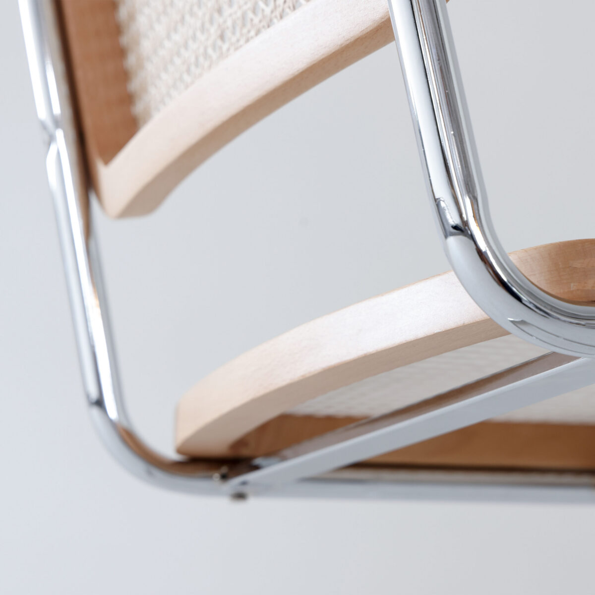 Natural Frame_Chrome_B2 Close-up of a modern chair showing a chrome metal frame, light wood seat and backrest, and woven cane detailing. The background is plain and light-coloured.