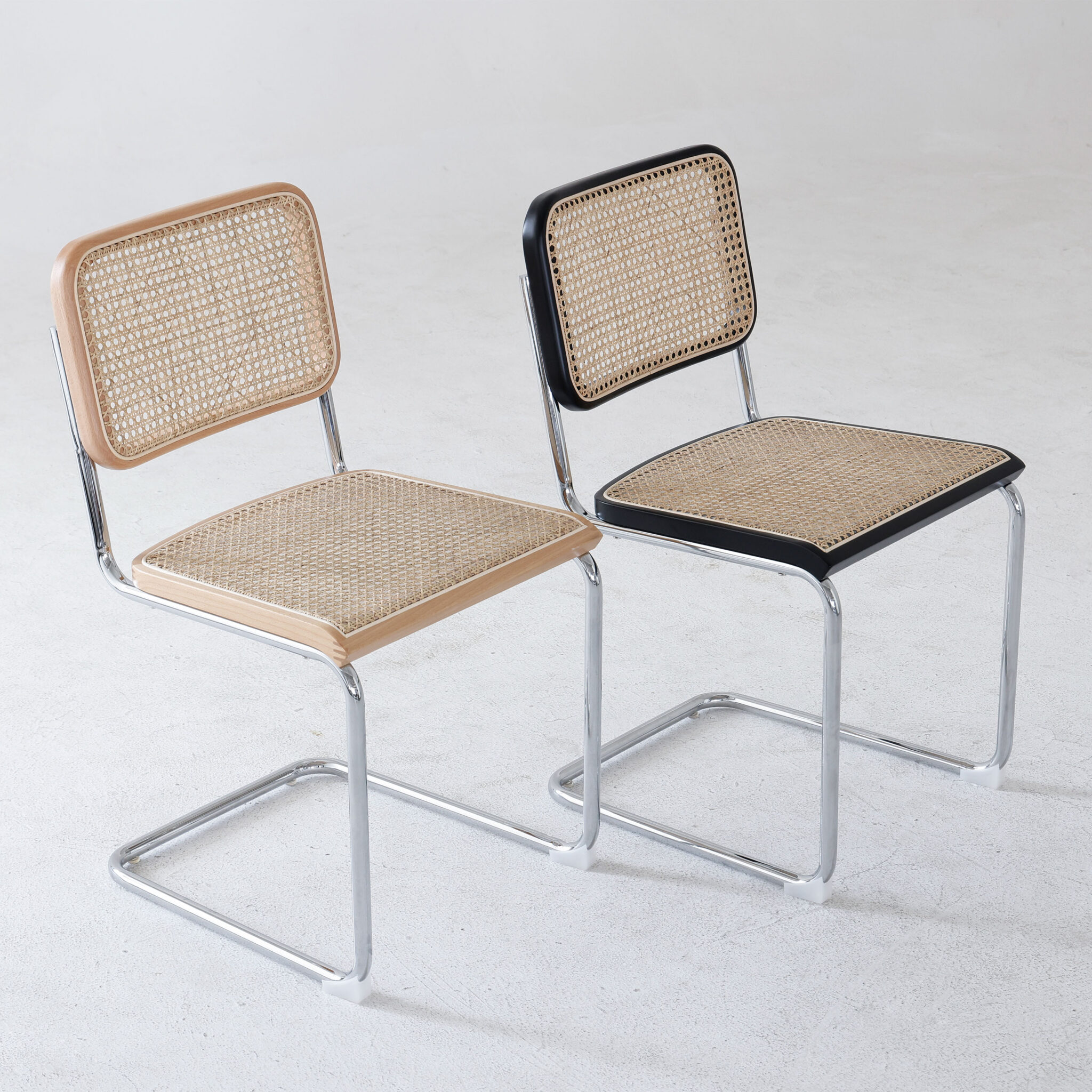 Two modern chairs with chrome cantilever frames, rattan seat and backrests. One chair has natural wood edges, the other has black edges. Both are set on a plain light-coloured floor with a neutral background.