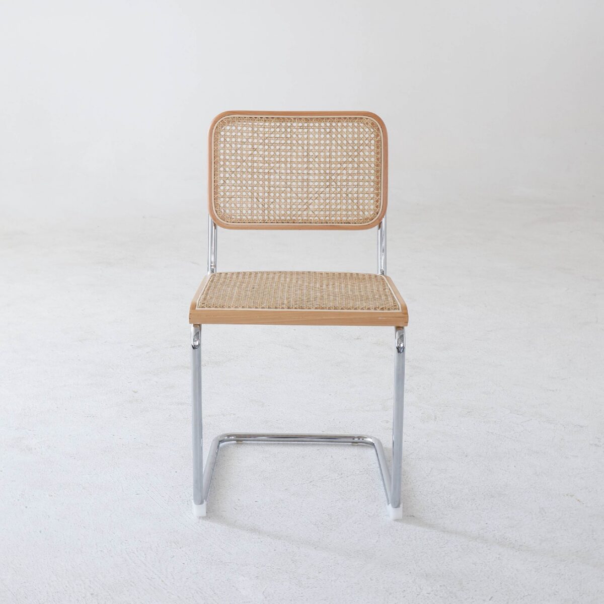Natural Frame_Chrome_B4 A chair with a chrome metal frame, a woven cane seat and backrest, and light wood accents, placed on a plain white floor against a white wall.