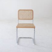 A chair with a chrome metal frame, a woven cane seat and backrest, and light wood accents, placed on a plain white floor against a white wall.