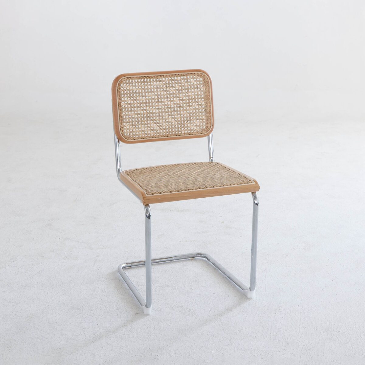 Natural Frame_Chrome_B5 A minimalist chair with a metal frame, a woven cane backrest, and a matching cane seat, placed on a plain white floor against a white wall.