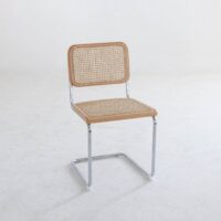 A minimalist chair with a metal frame, a woven cane backrest, and a matching cane seat, placed on a plain white floor against a white wall.