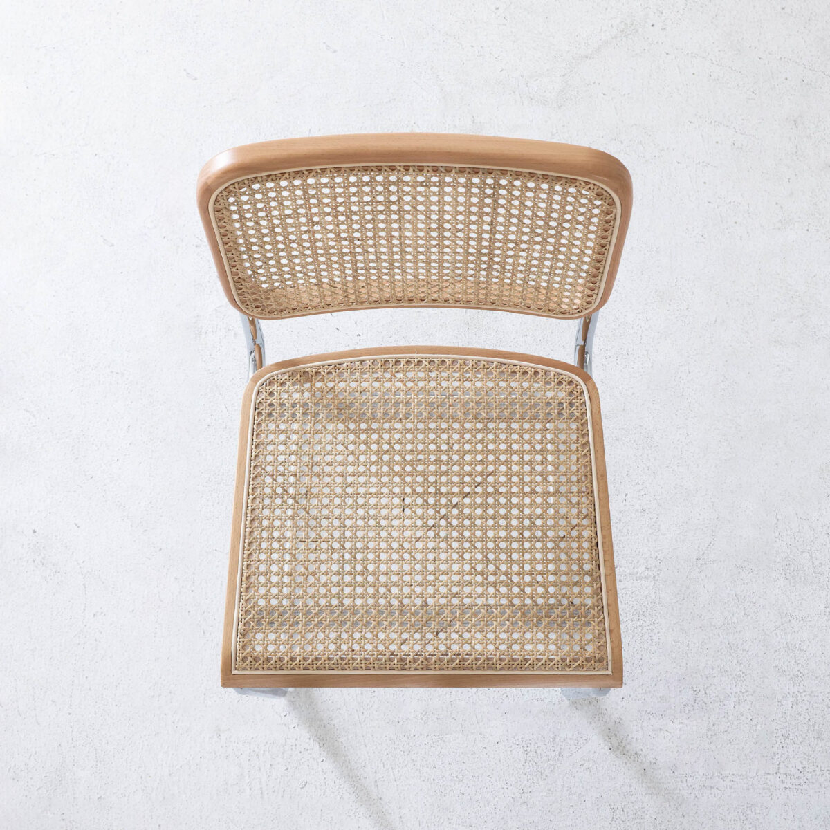 Natural Frame_Chrome_B7 A top-down view of a wooden chair with a woven cane seat and backrest, placed on a light, textured floor. The chair has a simple and minimalist design.