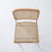 A top-down view of a wooden chair with a woven cane seat and backrest, placed on a light, textured floor. The chair has a simple and minimalist design.