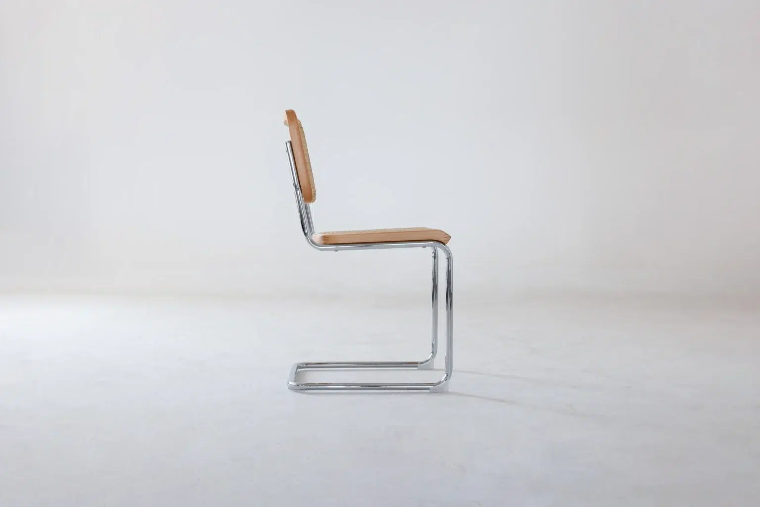 Side view A minimalist cantilever chair with a chrome tubular metal frame and light tan wooden seat and backrest, shown in profile against a plain white background.