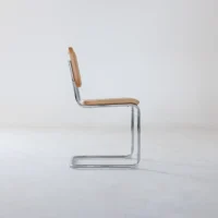 A minimalist cantilever chair with a chrome tubular metal frame and light tan wooden seat and backrest, shown in profile against a plain white background.