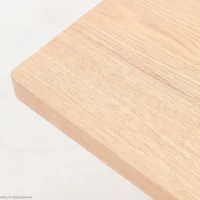 A close-up of the corner of a light-coloured wooden tabletop, showing its smooth surface and natural wood grain pattern against a pale background.
