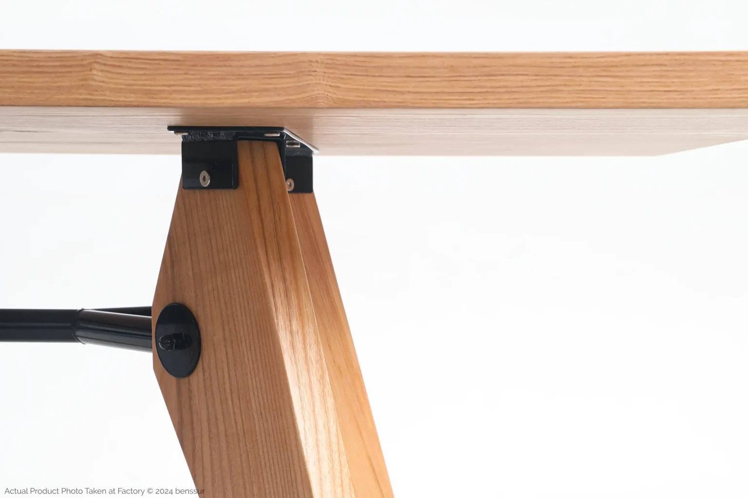 Jean Prouvé_S.A.M No.502 Table_Replica_Natural_Ash_Detail_2 Close-up of a wooden table leg attached to the tabletop with black metal brackets. The wood has a light, natural finish, and the background is plain white.