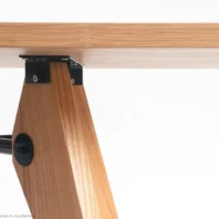 Close-up of a wooden table leg attached to the tabletop with black metal brackets. The wood has a light, natural finish, and the background is plain white.
