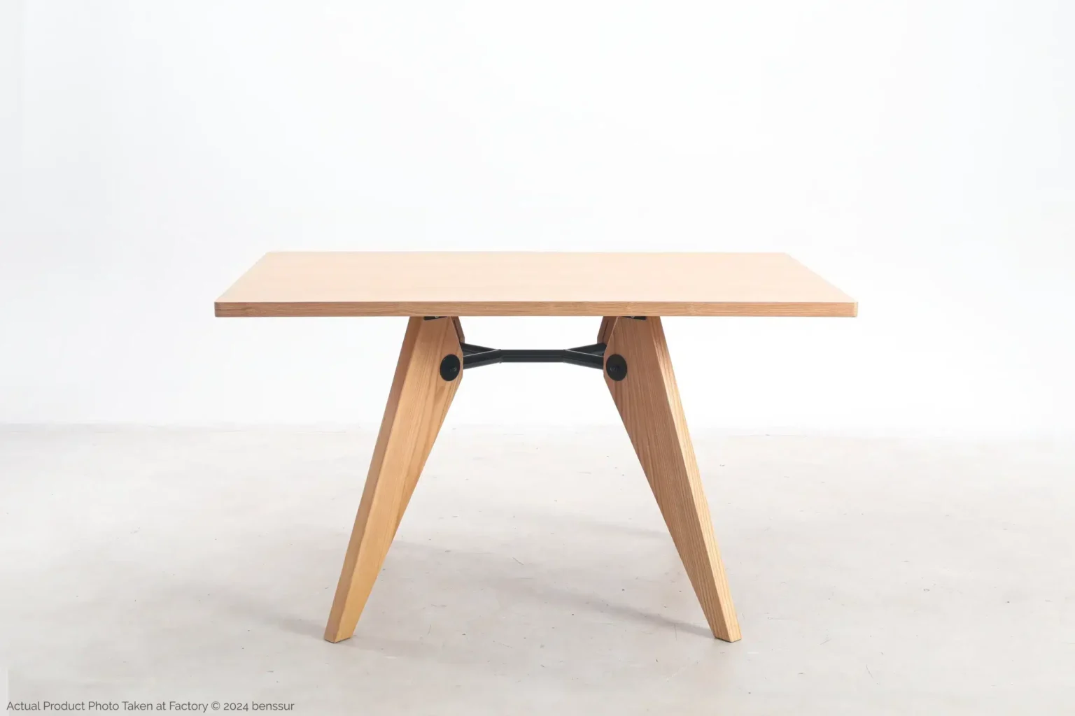 Jean Prouvé_S.A.M No.502 Table_Replica_Natural_Ash_Front A minimalist wooden table with a rectangular top and two V-shaped legs connected by a black metal support bar, set against a plain white background.