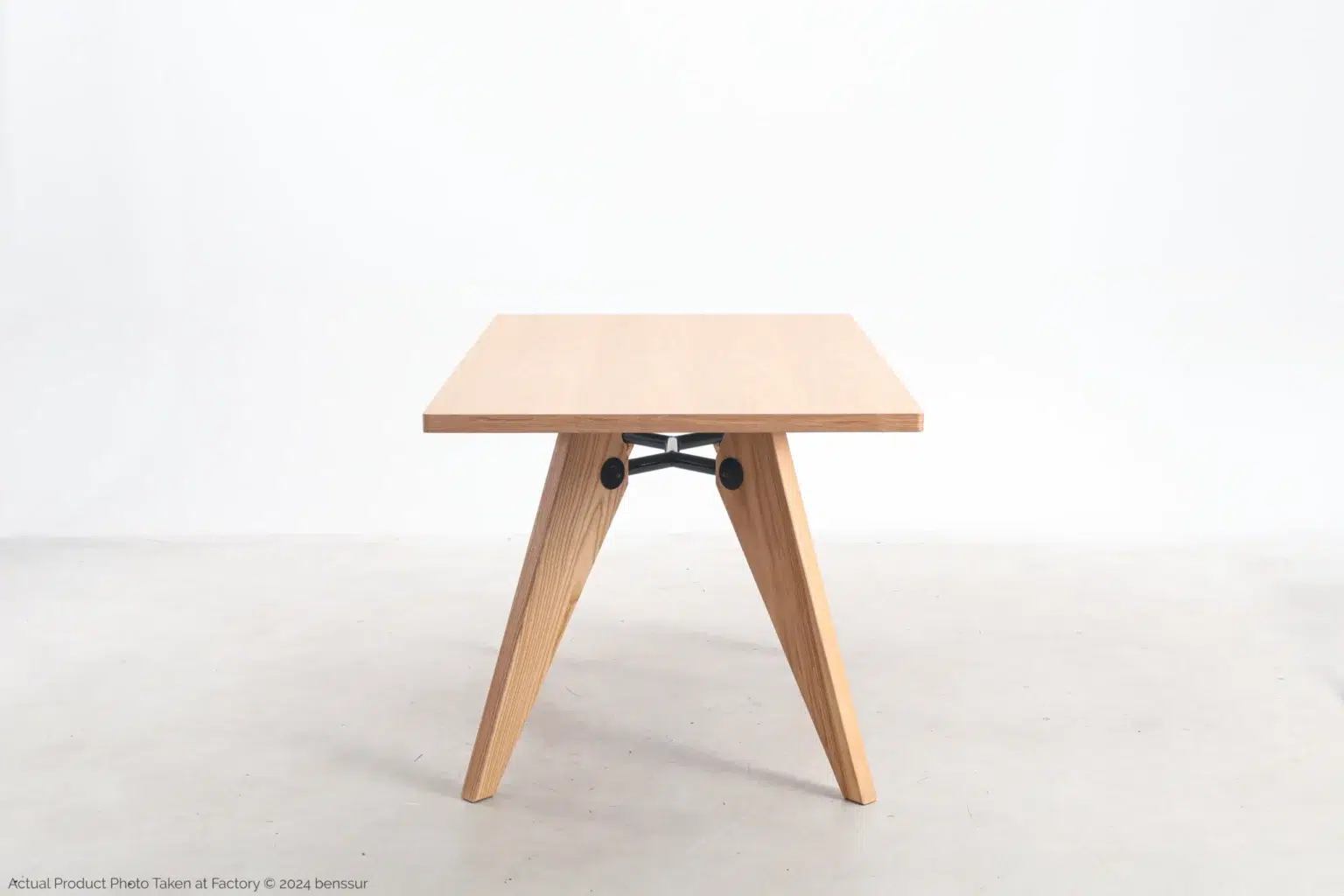 Jean Prouvé_S.A.M No.502 Table_Replica_Natural_Ash_Side A minimalist wooden table with a square top and angled, V-shaped legs, connected by a black metal support bar, set against a plain white background.