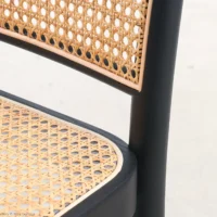 Close-up of a chair with a woven cane seat and backrest, framed by a smooth, black wooden structure. The background is plain and light-coloured.