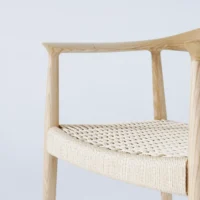 A close-up of a wooden chair with smooth, light wood and a woven seat, photographed against a plain light background. Only part of the backrest, armrest, and seat are visible.