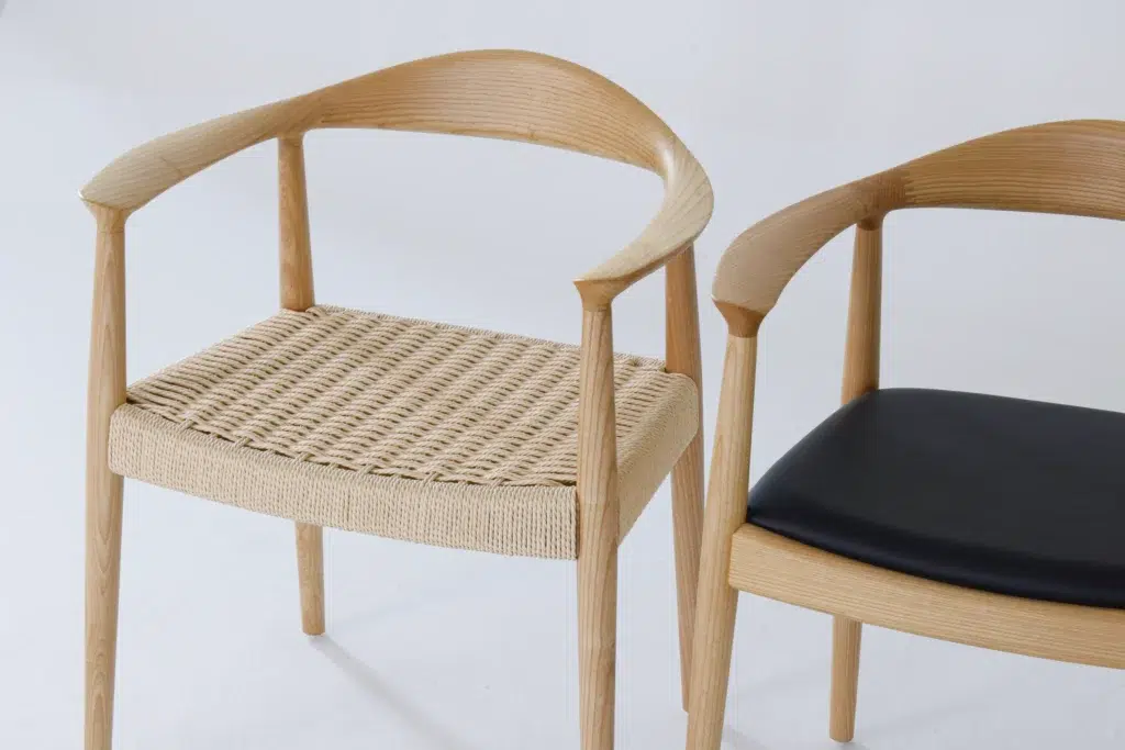 Two wooden chairs with curved backrests; one has a woven natural fibre seat, and the other has a smooth black cushion seat. Both chairs are on a plain white background.