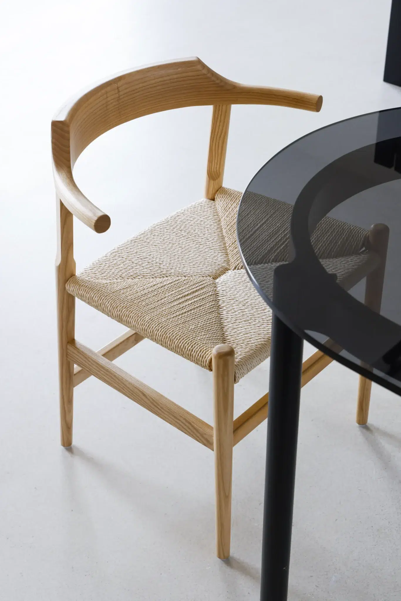 16 A wooden chair with a curved backrest and woven seat is positioned next to a round glass table on a light-coloured floor.