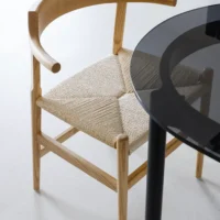 A wooden chair with a curved backrest and woven seat is positioned next to a round glass table on a light-coloured floor.