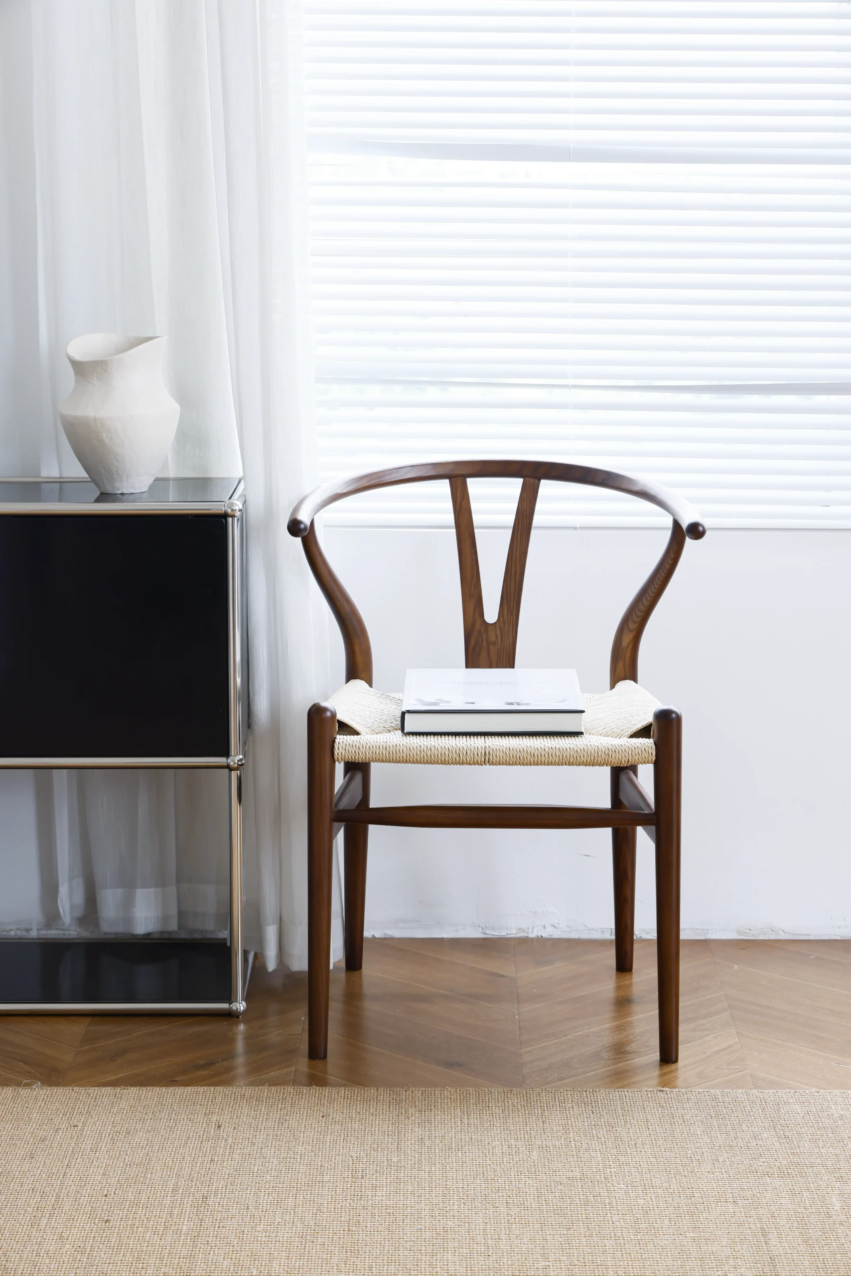 Hans Wegner's Wishbone Chair in dark brown, placed near a window with a book resting on the seat, creating a cosy and stylish scene that highlights the chair's iconic design and timeless elegance in a relaxed setting.