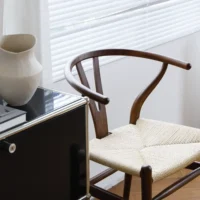 A wooden chair with a woven seat sits beside a glossy black cupboard topped with books and a cream-coloured ceramic vase, near a window with white blinds and sheer curtains.