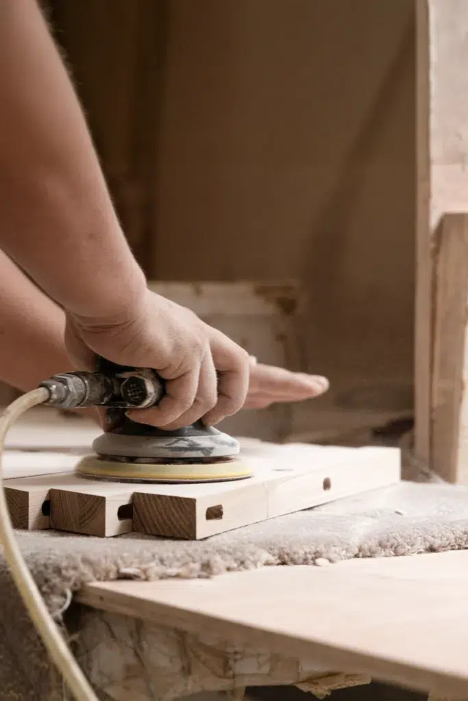 A person uses an electric sander to smooth the surface of a light-coloured wooden board, creating dust in a workshop setting.