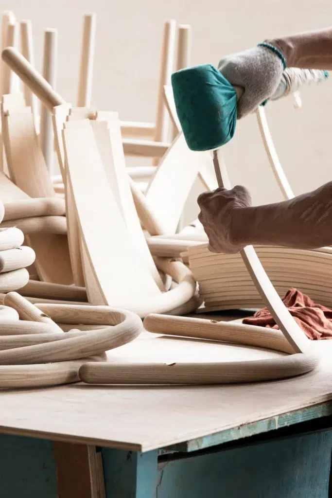 A person wearing a glove shapes or assembles light wooden chair parts on a workbench, surrounded by curved and straight wooden pieces in a workshop setting.