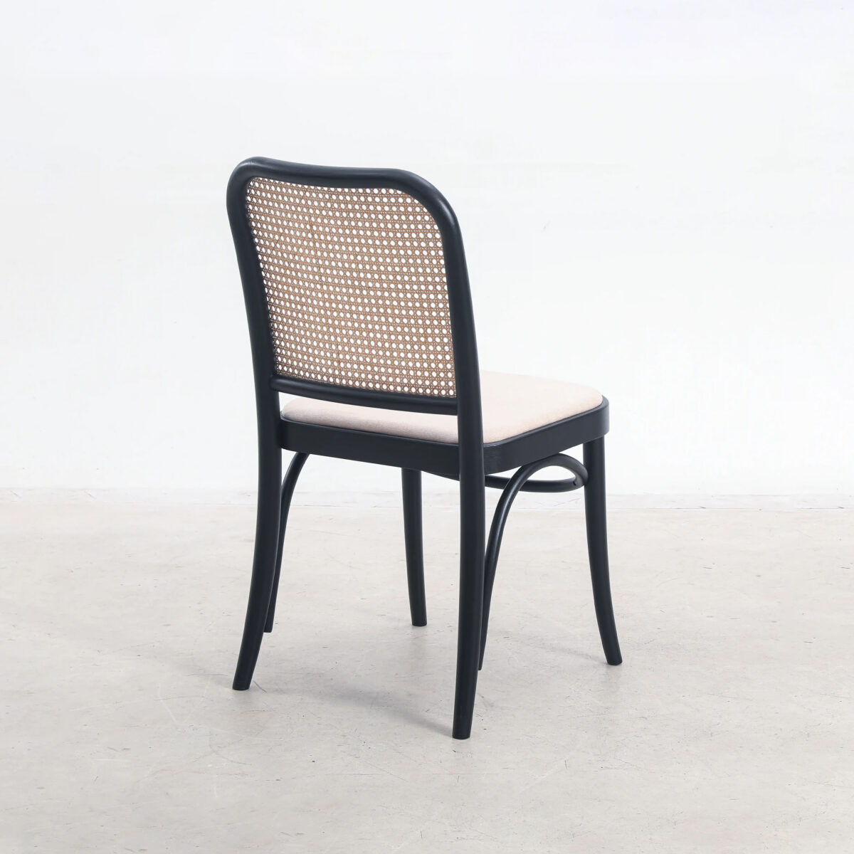 Fabric_B1 A black wooden chair with a curved back and a woven rattan backrest, viewed from behind, sits on a light-coloured floor against a plain white background.