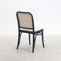 A black wooden chair with a curved back and a woven rattan backrest, viewed from behind, sits on a light-coloured floor against a plain white background.