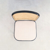 Top view of a chair with a light beige cushioned seat and a curved woven rattan backrest, framed in black, placed on a light grey, textured floor.