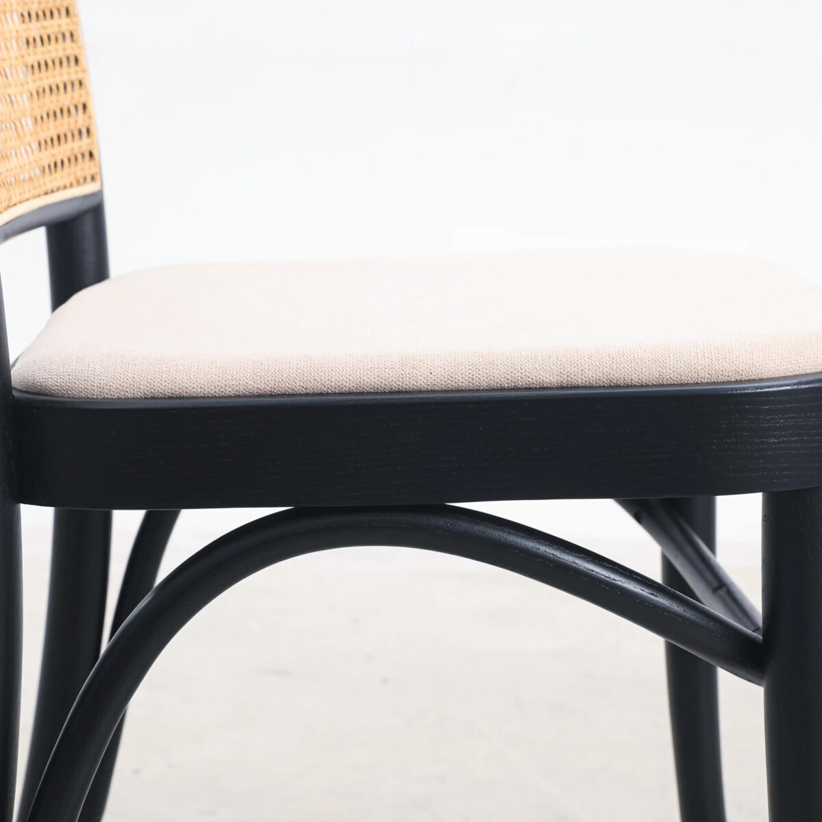 Fabric_B4 Side view of a chair with a light beige cushioned seat, black wooden frame, and curved support bars beneath the seat. The chair’s backrest partially features a woven pattern. Background is plain and light-coloured.