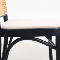 Side view of a chair with a light beige cushioned seat, black wooden frame, and curved support bars beneath the seat. The chair’s backrest partially features a woven pattern. Background is plain and light-coloured.