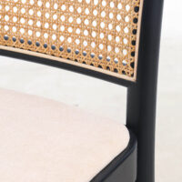 Close-up of a chair featuring a woven cane backrest, black wooden frame, and a light beige upholstered seat cushion.