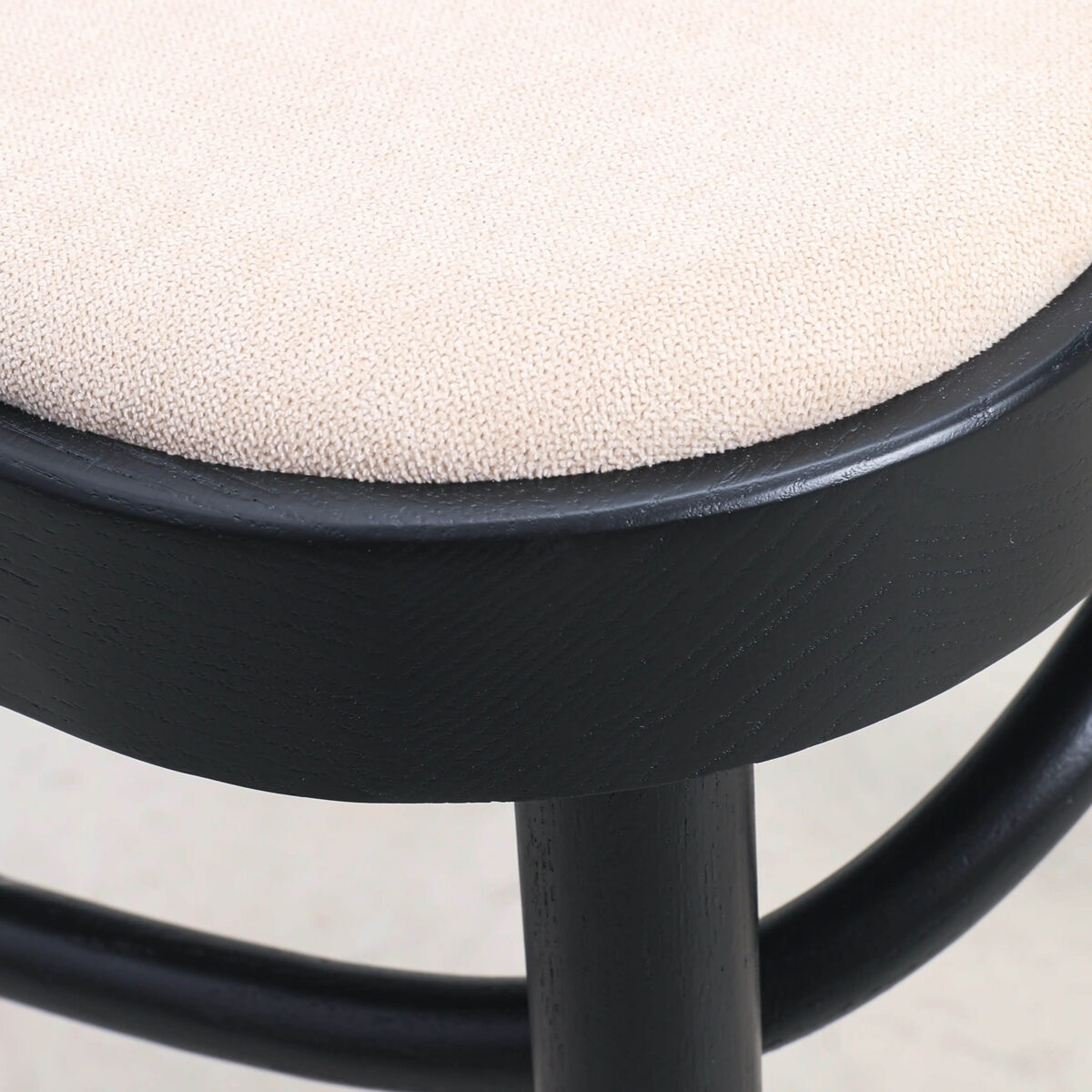 Fabric_B7 Close-up of a stool with a round, beige upholstered seat and a black wooden frame, showcasing the texture of the fabric and the smooth, curved legs.