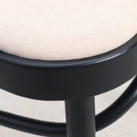 Close-up of a stool with a round, beige upholstered seat and a black wooden frame, showcasing the texture of the fabric and the smooth, curved legs.